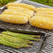 Two Outset non-stick metal grill trays with corn on the cob and asparagus cooking on a grill.