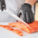 A person wearing black gloves uses a Mercer Culinary Sashimi Knife to cut a piece of salmon.