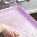 A hand washing a Baker's Mark purple aluminum sheet pan under a faucet.