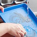 A person washing a Baker's Mark light blue aluminum tray with a sponge.
