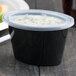 A black Pactiv oval plastic souffle container with a clear lid.