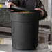 A man in a chef's uniform opening a large black round ingredient storage bin filled with colorful candies.