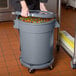 A woman using a gray mobile ingredient storage bin with lid in a school kitchen.