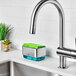 An OXO Good Grips soap dispensing sponge holder with a green sponge on the counter above a stainless steel sink.