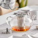 A silver kettle pouring liquid into a glass teapot with a stainless steel infuser.