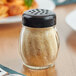 A Tablecraft glass cheese shaker with a black lid filled with brown powder on a table.
