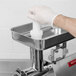 A hand wearing a glove uses a white plastic Avantco pusher to grind meat.