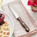 A Choice steak knife with a dark brown wood handle on a white napkin next to a plate of food.