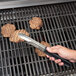 A hand using Vollrath Jacob's Pride tongs with a black handle to flip hamburgers on a grill.
