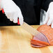 A person wearing white gloves uses a Mercer Culinary Millennia Colors Santoku knife with a red handle to cut ham.