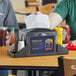 A OneUp by Choice Smoke Black Tabletop Napkin Dispenser with Condiment Caddy on a table with a person holding a plastic container of sauce.