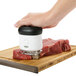 A hand using an OXO Good Grips meat tenderizer to stamp meat on a cutting board.