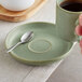 A hand holding an Acopa sage porcelain saucer with a cup of coffee and a spoon.
