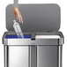 A woman's hand reaching for a plastic bottle in a simplehuman stainless steel sensor trash can.