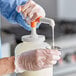 A gloved hand uses a Choice Maxi Pelican Condiment Pump to pour white liquid into a container.