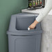 A hand opens a Lavex gray corner round push door trash can lid over a trash can.