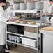 A chef in a white coat and gloves standing at a stainless steel Regency work table with shelves.