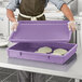 A person wearing a purple apron holding a purple rectangular plastic lid.