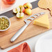 A cheese knife next to cheese and fruit on a cutting board.