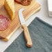 An Acopa stainless steel cheese knife with a wood handle on a cutting board next to cheese and meat.