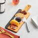 An Acacia wood and slate serving board with cheese, crackers, and blackberries on it, with a cheese knife and fork on a napkin.