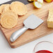 An Acopa stainless steel cheese knife on a wooden cutting board with cheese and crackers.
