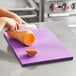 A person using a knife to cut a sweet potato on a purple Choice polyethylene cutting board.