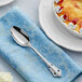 An Acopa stainless steel teaspoon on a blue napkin next to a dessert plate.