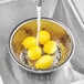 A stainless steel Chinese colander with lemons in it under running water.