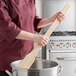 A woman in a chef's uniform uses a Choice wood paddle to stir a pot on a counter.