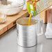 A person pouring orange juice from a metal container into a Choice aluminum measuring cup.