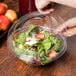 A person putting salad into a Fineline plastic bowl with a clear dome lid.