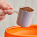 A hand using a long-handled polypropylene scoop to pour brown granules into a container.