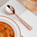 A Visions rose gold plastic soup spoon with a rose design on it sitting on a plate.