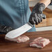 A person in black gloves using a Schraf Butcher Knife with a TPRgrip handle to cut meat on a wooden surface.