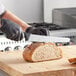 A person in black gloves uses a Schraf serrated bread knife to cut a loaf of bread on a wooden cutting board.