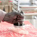 A person in black gloves using an Omcan stainless steel meat tenderizer on a piece of meat on a cutting board.