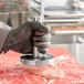 A person in black gloves using an Omcan stainless steel meat tenderizer on a piece of meat on a counter.