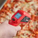A hand holding a red AvaTemp infrared thermometer over a pizza.