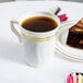A Fineline Silver Splendor ivory plastic coffee mug filled with brown liquid on a table with a slice of cake.