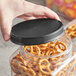 A hand holding a 110/400 black ribbed plastic cap over a plastic container of pretzels.