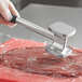A person uses a Choice 10" aluminum meat tenderizer with a rubber handle to hammer meat on a counter.