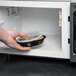 A hand putting a Pactiv Newspring VERSAtainer oval plastic container with food into a microwave.