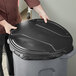 A man in a chef's uniform holding a black plastic lid over a Toter 44 gallon round trash can.