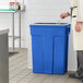 A person in a white towel putting a plastic bottle into a blue Toter Slimline trash can.