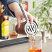 A person using a Franmara stainless steel cocktail strainer to pour orange liquid into a glass.