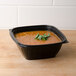 A Dart black square plastic bowl filled with soup and garnished with a sprig of parsley.