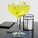A Libbey Super Margarita glass filled with a yellow drink and lime slices.