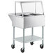 A stainless steel ServIt electric steam table on wheels with an angled glass sneeze guard.