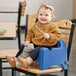 A baby sitting in a Carlisle blue booster seat on a chair holding a spoon.